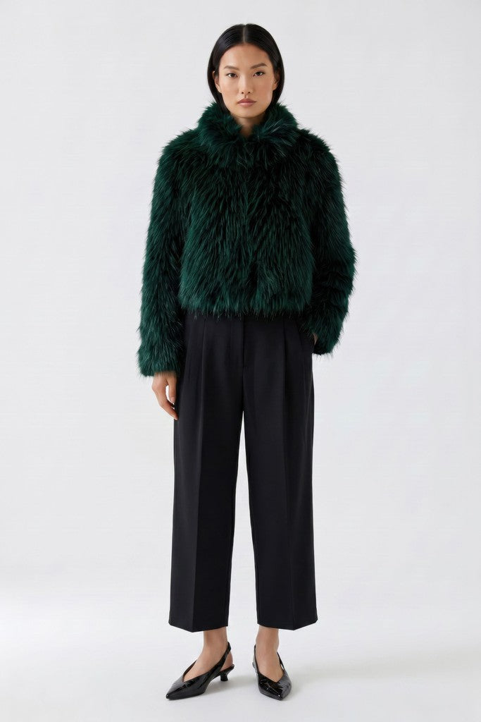 A model stands against a light gray studio background wearing a dark green textured faux-fur jacket with a high collar, black wide-leg cropped trousers, and black pointed flats.