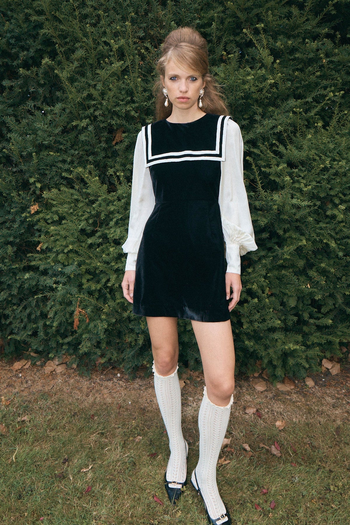 A woman stands outdoors in front of a dense green hedge, wearing a black velvet mini dress with white trim and sheer white long sleeves, paired with white knee-high socks and black-and-white shoes.