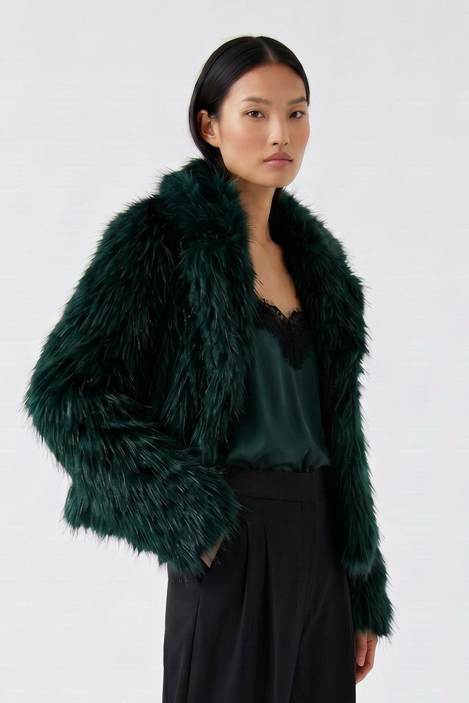 A model wearing a dark forest green faux fur jacket over a matching dark green satin top and black trousers, photographed against a clean white studio background.