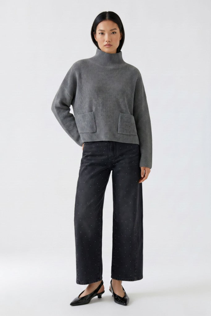 A model stands against a light gray studio background wearing a gray ribbed turtleneck sweater with front patch pockets, dand grey sparkle denim barrel jeans and black pointed flats.