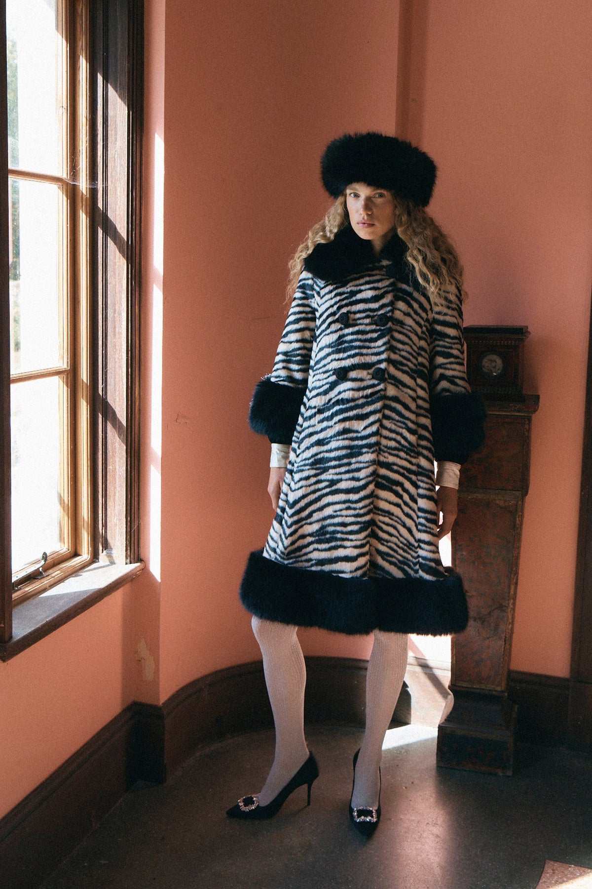 A woman stands in a sunlit room wearing a black-and-white zebra-pattern coat with dark fur trim, a matching fur hat, white tights, and black heels.