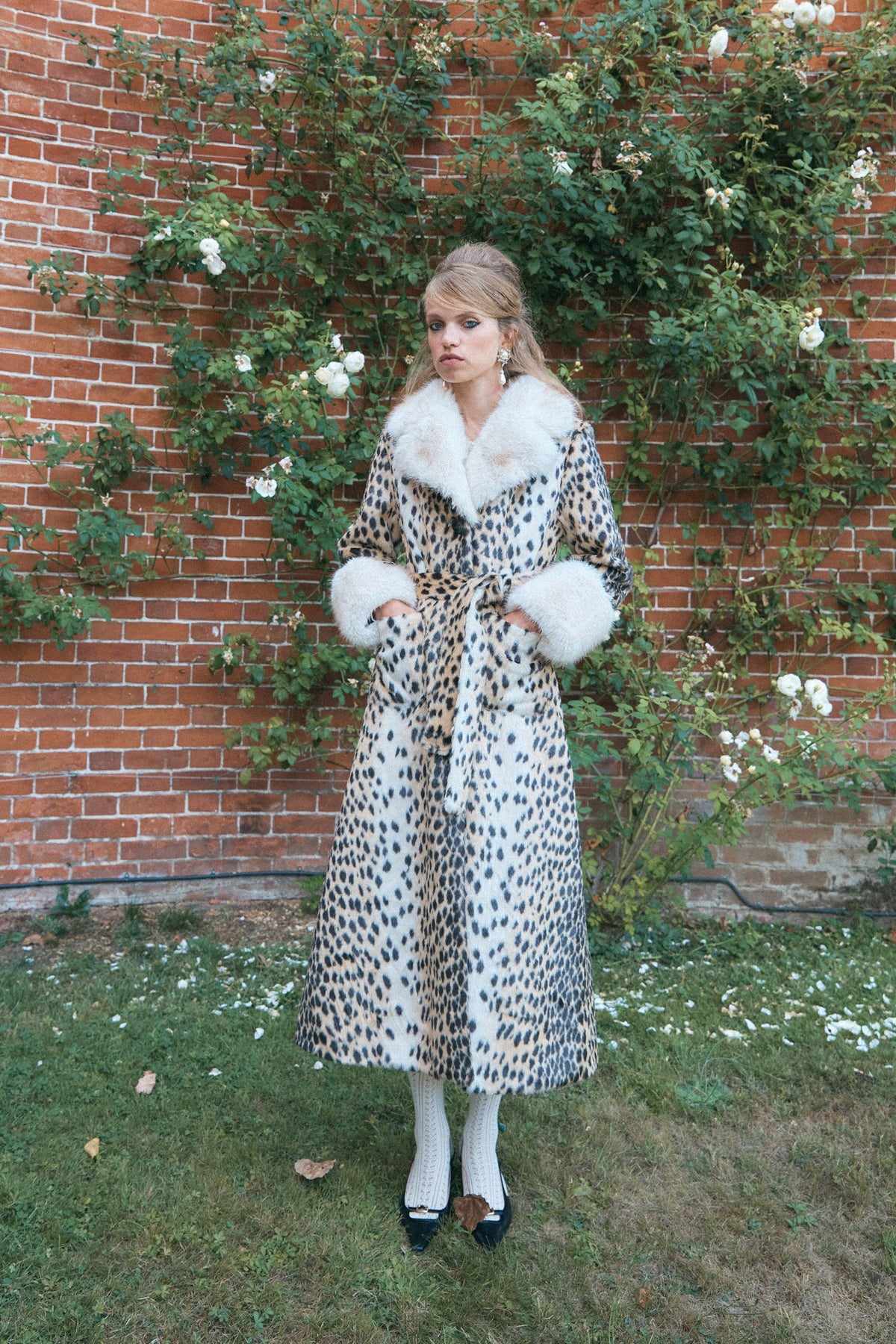 A woman wearing a long leopard-print coat with a large white fur collar and cuffs, white tights, and black shoes.