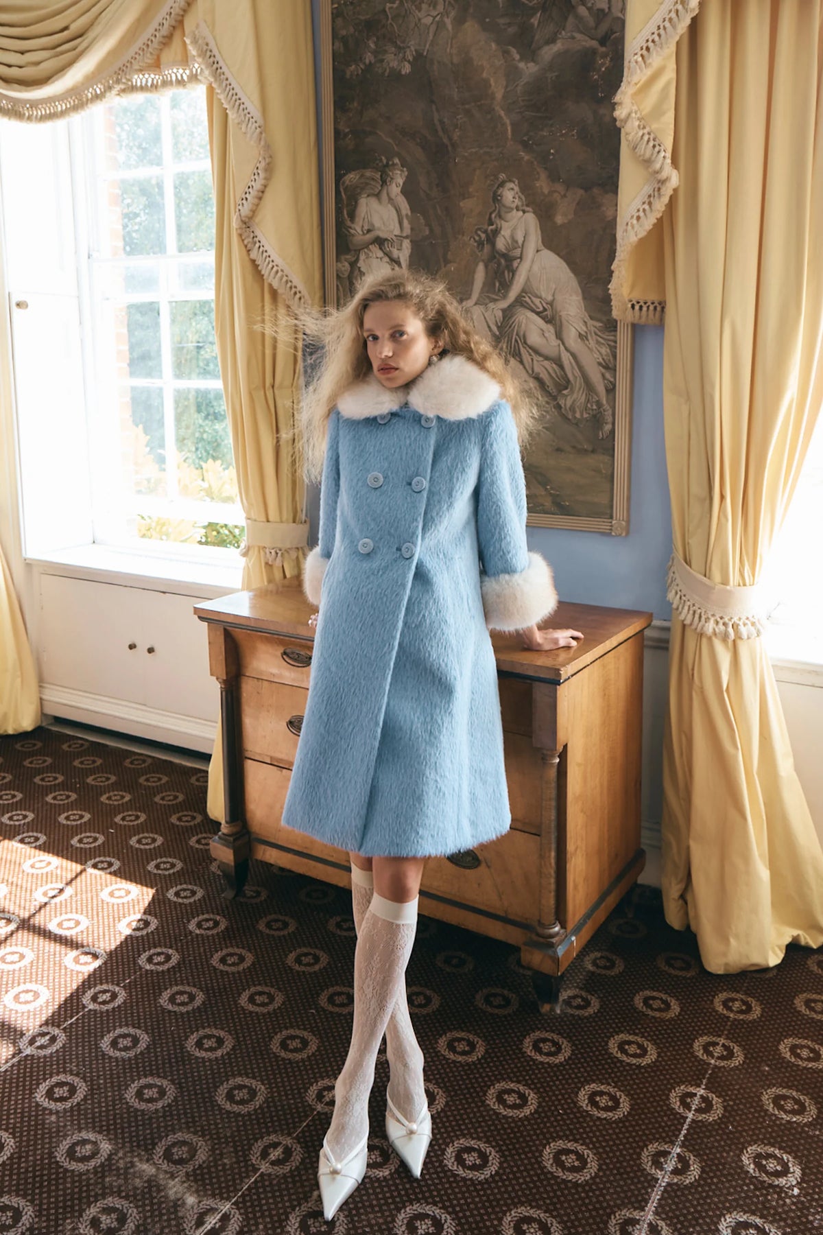 A woman stands in a vintage room wearing a light blue textured coat with a white fur collar and cuffs, white thigh-high stockings, and white pointed shoes. She is posed in front of a wooden chest and a large framed painting, with yellow curtains and a patterned carpet visible.