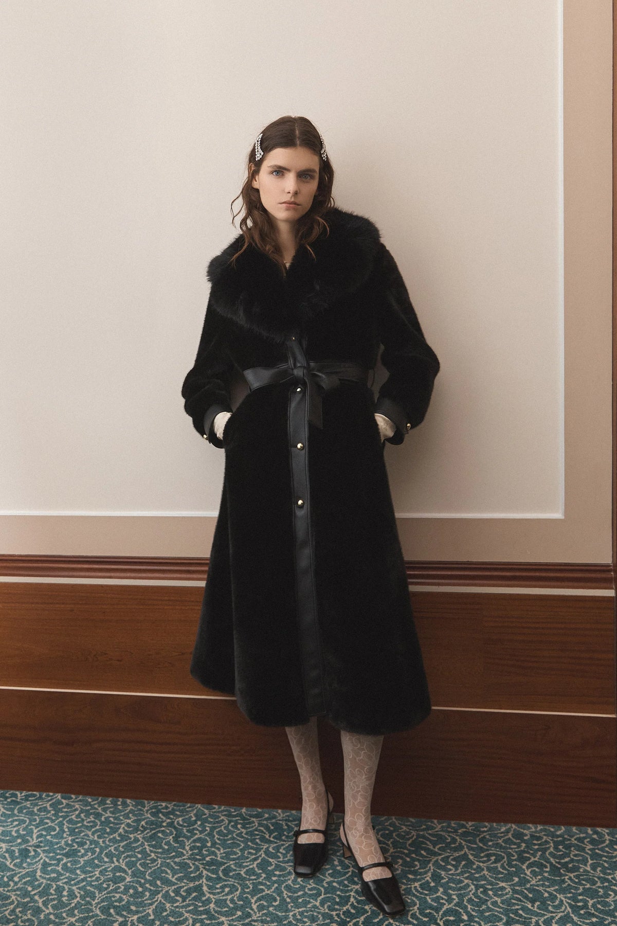A model stands indoors wearing a long black fur coat with a dark satin waist belt and front buttons, paired with patterned tights and black shoes, against a beige wall with wood trim.